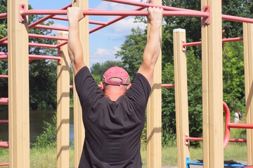 one male athlete in a black shirt pulls himself up on a red sports horizontal bar on the street on a sports ground against a blue sky
