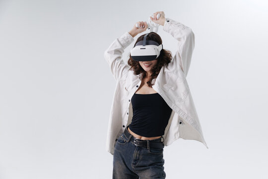 Young hispanic female with curly hair enjoying virtual reality headset experience, online video game of dance on white studio background