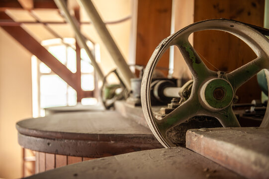 Innenansicht einer historischen Getreidem&uuml;hle mit Laufrad und Transmissionsriemen auf Holzbottich in im Gegenlicht vor unscharfem Hintergrund mit Bogenfenster.
