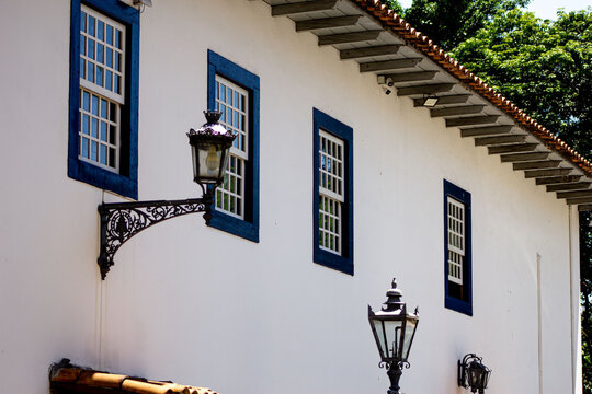 Parede com janelas de forma geom&eacute;trica, telhado com telha de barro, l&acirc;mpada arandela estilo colonial. 