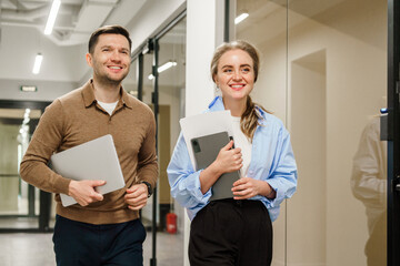 Two people walk together in a modern office carrying laptops and documents during work hours in a bright environment