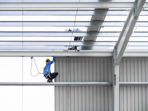 Construction workers wearing safety harnesses installing metal wall panels on a large steel frame industrial building.