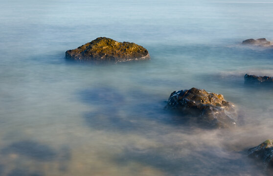 Close-up Long Exposure of Sea Waves Crashing Over Stones. A detailed long exposure shot showing the silky movement of water around dark rocks. The mist-like texture of the water creates a calm and eth