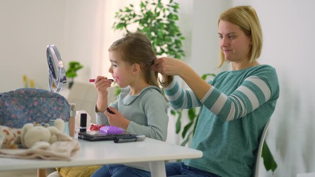 Mother braiding her daughter's hair while she is applying lipstick