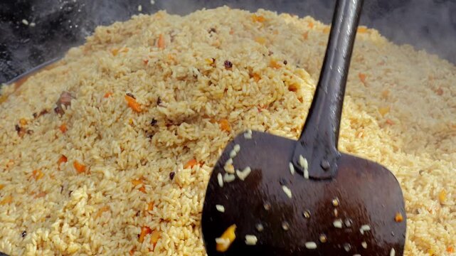 Steaming pilaf rice being stirred with ladle, fragrant grains with carrot and spice rising steam from giant wok at outdoor food festival, slow rhythmic mixing by vendor