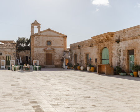 View of sun-drenched stone buildings, including a church with a bell tower, line a wide-open square, creating a warm, textured scene, Marzamemi, Sicily, Italy.