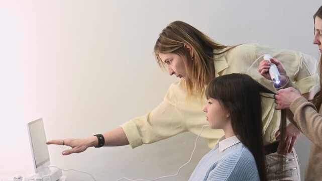 hairdresser examines a young woman's long, natural chestnut hair. A trichologist diagnoses condition of her hair and scalp. Two professional hairdressers trichologists and client looking on screen