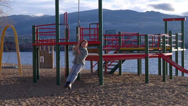 Young girl in cold weather clothing riding on a zipline located at a lakeside playground, coming towards the camera with a big smile.