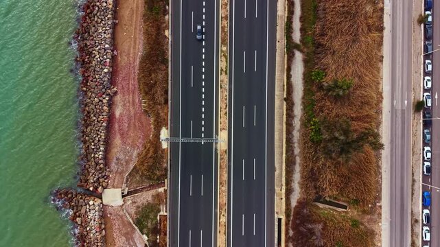 Vehicles move along a highway next to the water. The view shows a stretch of road with traffic flowing in two directions under clear skies. The shoreline is visible along one side.
