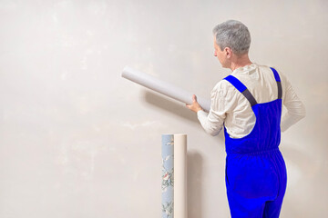 The gray-haired man in a blue jumpsuit is holding roll of wallpaper. Wallpaper sticker.