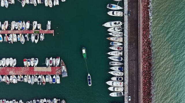 Boats are docked in neat rows at a marina. One boat is in the water and is being towed away. The sun lights up the scene in the early afternoon near the shore.