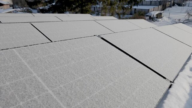 Close-up of rooftop solar panels covered with frost and light snow on a cold winter day. Residential neighborhood and trees visible in the background. Natural light, shallow depth of field