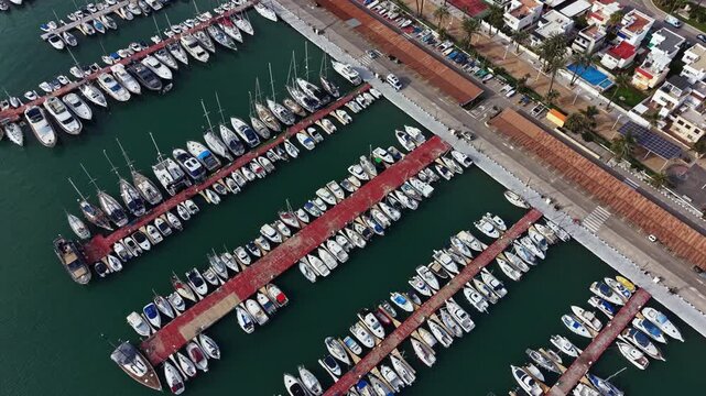 Many boats are lined up in a marina, with docks separating them. Nearby, there are houses and palm trees, showing daily life by the water.