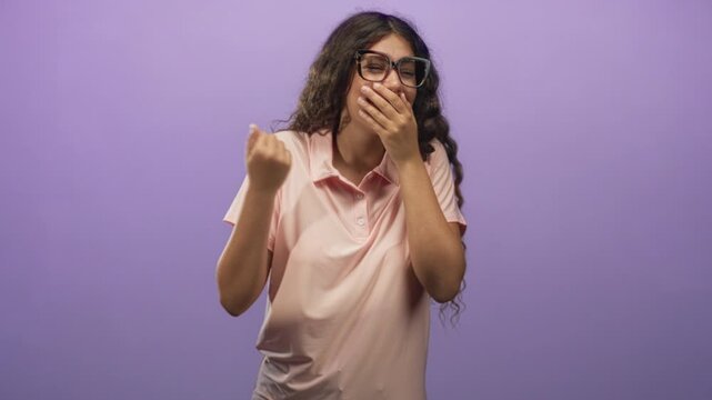 Hispanic teenage woman in pink polo covers mouth with hand while laughing and pumping fist in studio; playful amusement.
