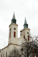 Obraz premium Twin church towers with clocks rising above winter trees in Sombor, Serbia. Symmetrical European landmark and cultural heritage concept.