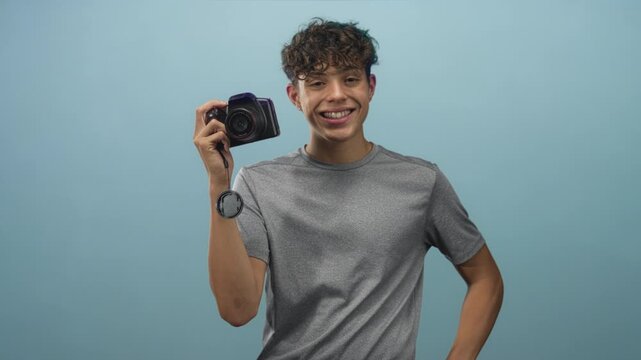 Man holding dslr camera with dangling lens cap in studio portrait, smiling and posing with hand on hip; confident youth.