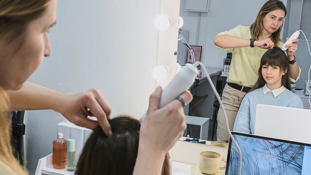 Trichologist examines hair and scalp of young woman in  salon using trichoscope and dermatoscope near large mirror. Salon interior professional occupation of hairdresser