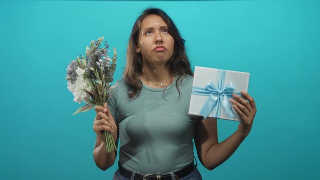 Woman holding bouquet and gift box with hands, pouting and looking up in turquoise studio; disappointment solitude contemplation.