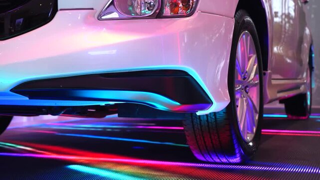 Low angle view of a modern white car's front bumper, wheel, and headlight standing on an interactive led floor with vibrant, colorful light lines reflecting on its shiny surface