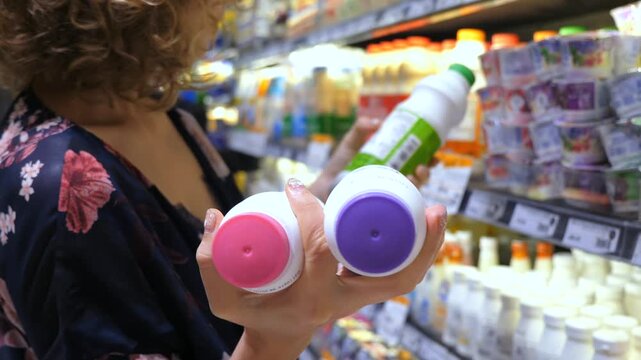 Young woman standing in a supermarket aisle holding two bottles of drinkable yogurt, carefully choosing dairy products for a healthy diet and comparing ingredients on the labels