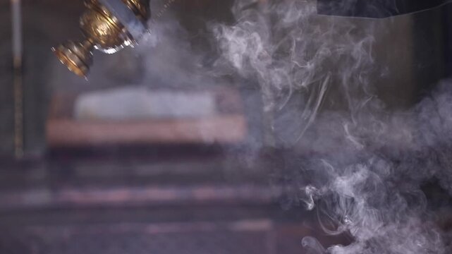 Close-up of a priest swinging a smoking censer during liturgy. The background features a blurred altar with candles, creating a solemn, spiritual atmosphere inside the traditional church.

