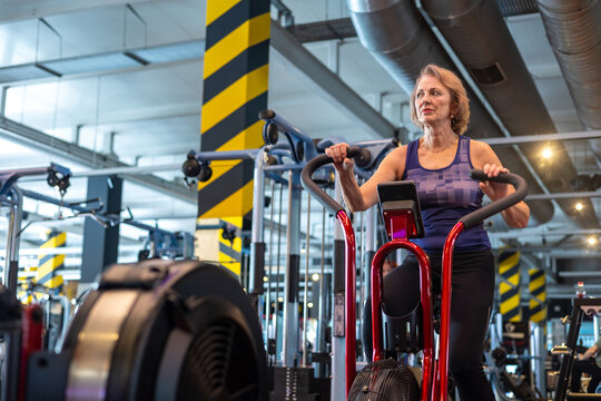 Senior woman riding air bike in modern gym, smiling during cardio workout, active lifestyle and healthy aging. cardiovascular health and redefining physical capability after retirement. postmenopause,