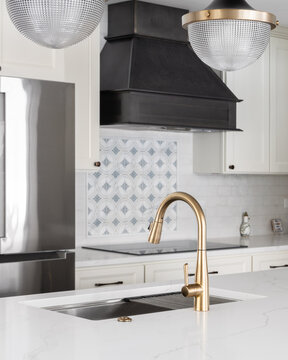 A gold kitchen faucet detail with black and gold lights hanging over an island, cream cabinets, and a pattern backsplash under a dark range hood.