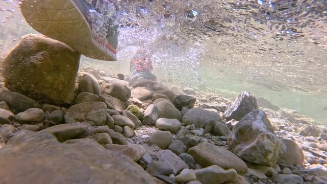 UNDERWATER, CLOSE UP: Unrecognizable woman in sneakers walks along a shallow riverbed. Bubbles swirl over smooth stones in clear fresh water as she wades through cold mountain stream on a sunny day.