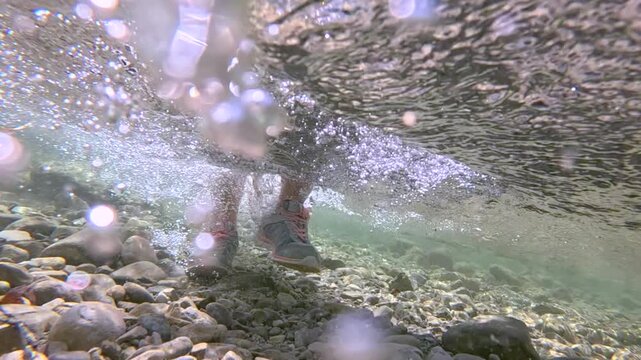 UNDERWATER, CLOSE UP: Unknown female hiker in sports shoes wades through a clear mountain stream on a sunny day. Air bubbles and turbulence swirl over gravel bed as sunlight filters through surface.