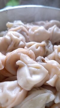 Close-up view of freshly cooked pelmeni dumplings piled in a white bowl. Hot steam rises from the moist dough pockets filled with meat, highlighting the soft appetizing texture and heat.