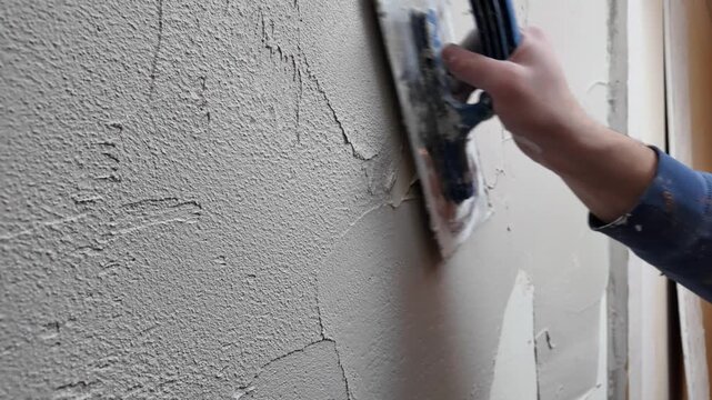 A person works on a wall by scraping off old plaster and smoothing new plaster on top. The activity takes place in a construction space during daytime.