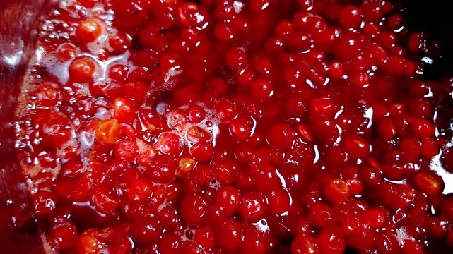Red berries boil and simmer in a dark pot, creating a thick, vibrant syrup for homemade jam. A close-up shot captures the bubbling texture and rich red color of the cooking fruit.