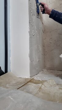 A man is smoothing a plastered wall with a trowel in a building area. He works on the corners and edges to create a flat surface.