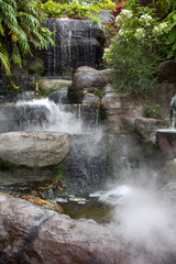 Naklejka premium Close up of a lush, tranquil water feature within a temple garden. Small waterfalls cascade over dark rocks into a misty pool, surrounded by vibrant green ferns and white hydrangeas.