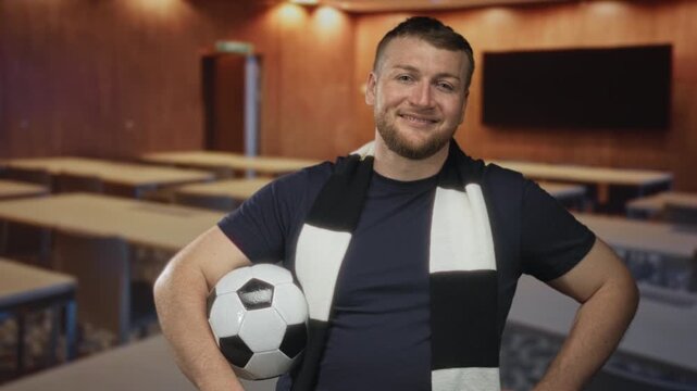 Man holding soccer ball and wearing black and white scarf in classroom building, smiling with hand on hip; pride and support.