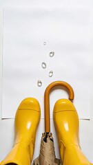 Yellow rain boots standing beside orange umbrella with falling raindrops on white background. Spring equinox weather symbol and seasonal renewal concept. Flat lay for greeting card design. portrait