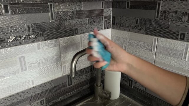 Hand squeezing dishwashing sponge with detergent foam near kitchen sink faucet closeup cleaning routine hygiene concept household chores washing dishes domestic sanitation lifestyle