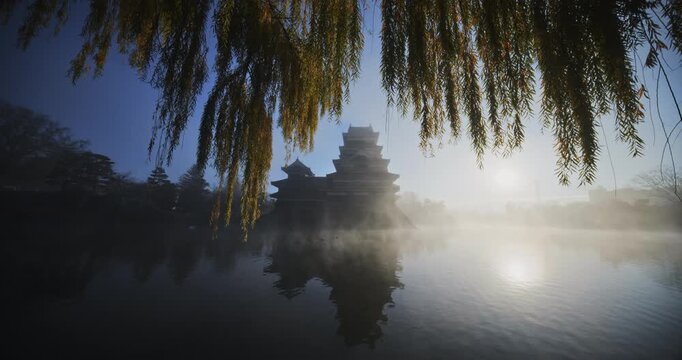 Rising sun shining through haze of pond near ancient 16th century Japanese castle - wide shot