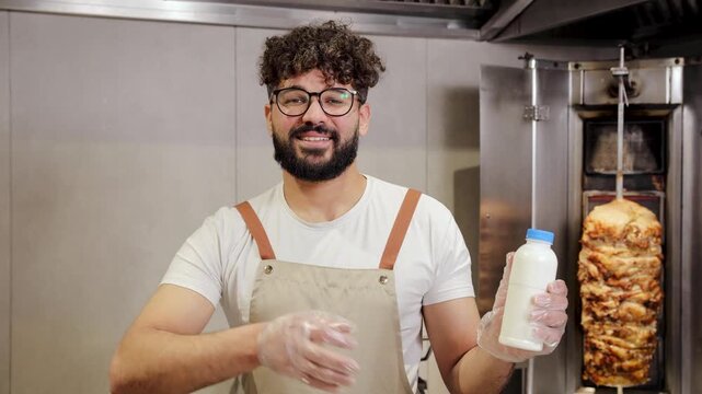 Chef, wearing an apron and gloves, showing a bottle of ayran as the best drink for kebab while standing by a vertical rotisserie in a restaurant kitchen