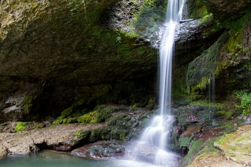 Obraz premium Lush waterfall flowing from a mossy rock cave into a small emerald pond.