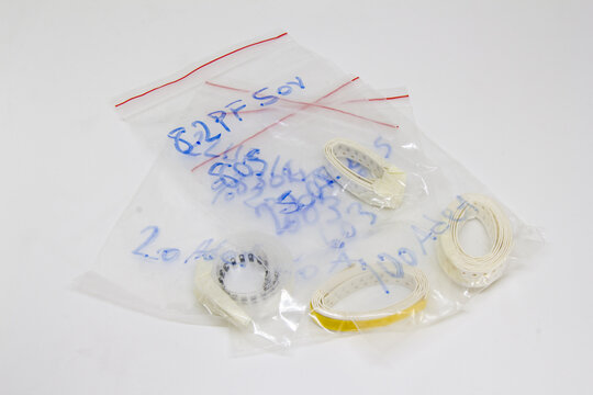 High angle close up view of SMD ceramic capacitor components on paper cut tape strips packaged inside clear ziplock plastic bags with handwritten blue marker notes on a white background
