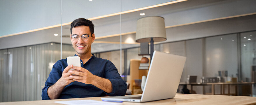 Smiling busy professional Hispanic business man sitting in office holding mobile cellphone. Young happy businessman entrepreneur using smartphone looking at cell phone technology device at work.
