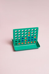 A classic four in a row strategy game with a green board and orange and blue discs. Minimalist studio shot on a pastel pink background. Concept of play, logic, competition, and nostalgia.