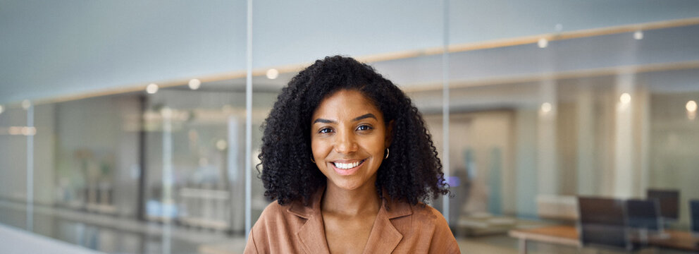 Happy confident young African American business woman, entrepreneur or employee standing in office. Smiling professional businesswoman with curly hair looking at camera at work, headshot portrait.