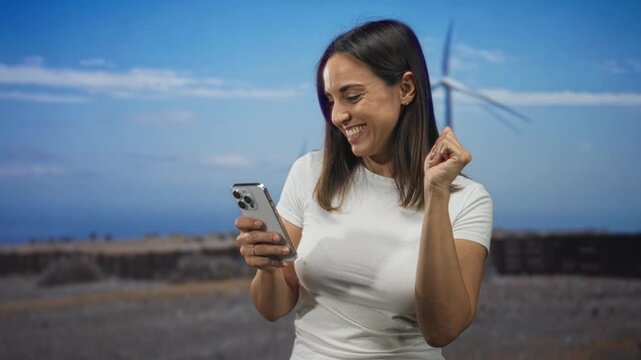 Woman checking smartphone with small fist pump gesture at wind farm field while smiling and holding phone; joy connection.
