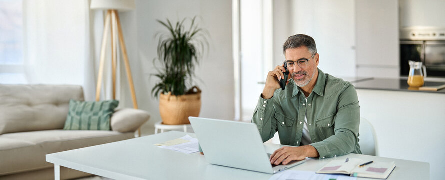 Happy busy middle aged mature man senior entrepreneur talking on mobile phone looking at laptop using computer calling on cellphone working at home sitting at kitchen table.