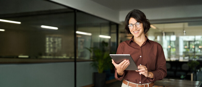 Smiling young Hispanic professional business woman wearing glasses using tablet standing in office. Happy businesswoman company manager, female entrepreneur or employee looking at camera. Portrait.