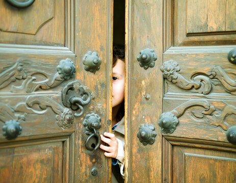 A curious child peeks through a narrow gap in a heavy, ornate wooden door. This evocative image symbolizes mystery and discovery, perfect for themes of storytelling, history, and secrets.
