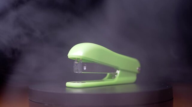A bright green office stapler rotates slowly on a black turntable while white smoke drifts through the dark air. Concept of office supplies, mysterious atmosphere, and dramatic product presentation