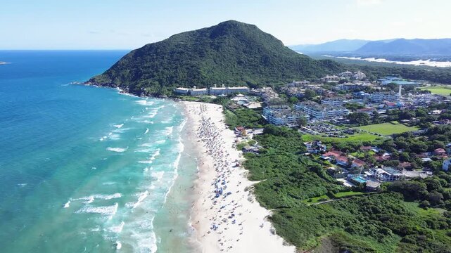Cinematic aerial panorama of Santinho Beach in Florianopolis, Brazil, featuring turquoise ocean waves, white sand shoreline and the lush green Morro das Aranhas mountain under a bright sky.
